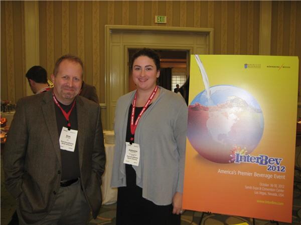 A man and a woman wearing badges stand beside an InterBev 2012 poster showing a globe, with dates October 16-18, 2012 and Las Vegas, Nevada.