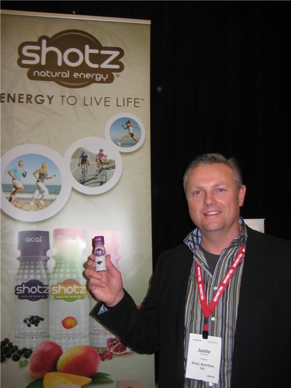 Jamie, President of Shotz Nutrition, Inc., holds a small Shotz bottle in front of a Shotz natural energy banner showing acai and other flavored bottles.
