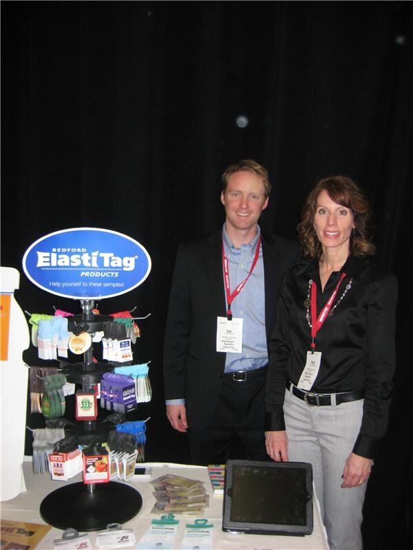 Jay stands beside a woman at a Bedford ElastiTag Products booth, with a spinning rack of colorful sample tags and a tablet on the table.