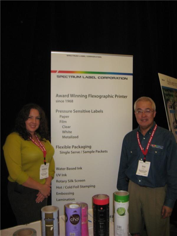Jerry, wearing a badge and red lanyard, stands at right beside a woman in a green top in front of a Spectrum Label Corporation banner, with rolls of printed labels on the table.