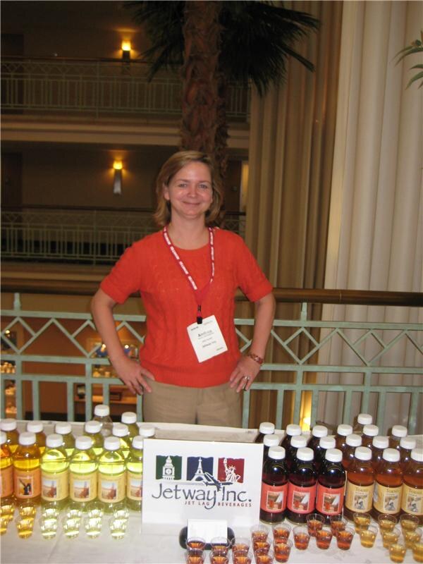 A smiling woman in an orange sweater with a badge stands behind a table of bottled teas and small sample cups, with a Jetway Inc. sign in front of her.