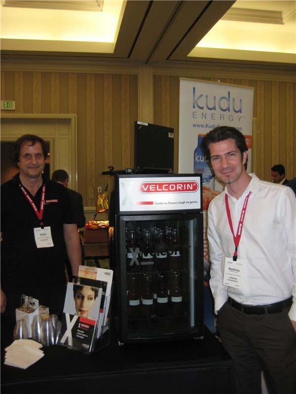 Matthias of LANXESS Corporation stands at right beside a Velcorin refrigerated display of bottles; another man in black stands at left, with a Kudu Energy banner behind.