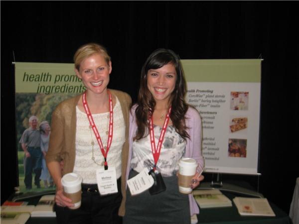 Melissa, left, and a second woman smile with coffee cups in front of a banner reading health promoting ingredients, both wearing red lanyards.