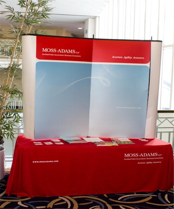 Moss-Adams LLP booth with a red and blue pop-up display and a red table cover, with brochures and cards on the table and a potted plant at left.