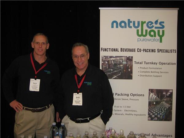 Two men in black shirts with lanyards and name badges stand beside a Nature's Way Purewater banner about functional beverage co-packing, with water bottles on the table in front.