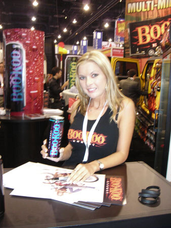 A smiling blonde woman in a black BooKoo tank top holds a can of BooKoo at a table with printed flyers, with other trade-show booths visible behind her.