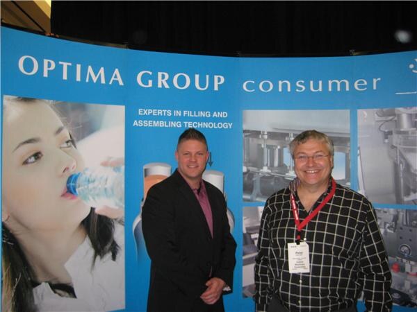 Two men stand in front of an Optima Group consumer backdrop about filling and assembling technology; Peter, on the right, wears a plaid shirt and a lanyard badge.