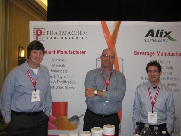 Three men in gray button-up shirts and lanyards stand in front of a Pharmachem Laboratories and Alix Technologies banner, with jars and bottles on the table below.