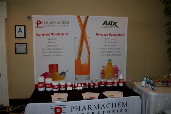 Booth with a Pharmachem Laboratories and Alix Technologies banner listing ingredient and beverage manufacturing, with product jars, bottles and brochures on a black table.