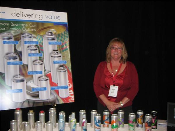 A woman in a red top with a lanyard stands beside a Rexam beverage can specification poster, with a row of aluminum cans and bottles on the table in front.