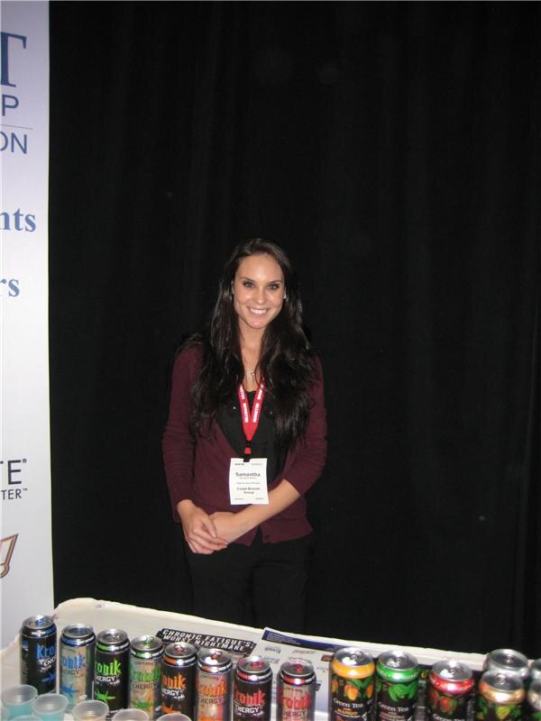 Samantha of Coast Brands Group smiles behind a table lined with Kronik Energy and green tea cans, wearing a red lanyard and maroon cardigan against a black curtain.