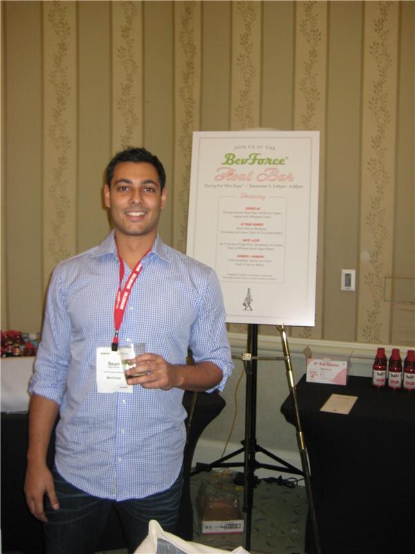 Sean, wearing a blue checked shirt and badge, smiles and holds a small cup in the foreground beside a BevForce Float Bar sign on an easel. Bottles sit on a table behind.