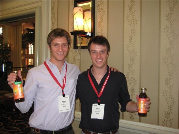 Tyler of Runa, left, and Dan of Runa, right, smile with arms around each other, each holding a bottle of Runa drink, in front of a wall with striped wallpaper.