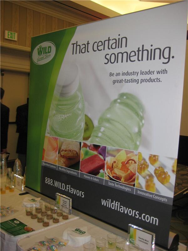 A WILD Flavors display reading "That certain something." with product images, beside a table with small sample cups, packets and signs including Vanilla Mint Lemonade.