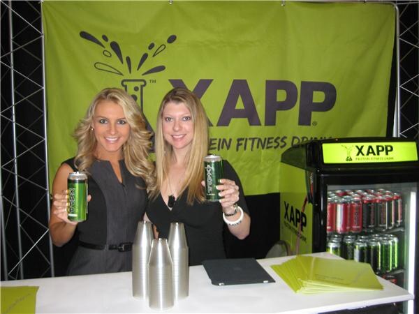 Two blonde women hold green XAPP cans behind a white counter, with a green XAPP banner behind them and a lit refrigerator of XAPP cans at right.