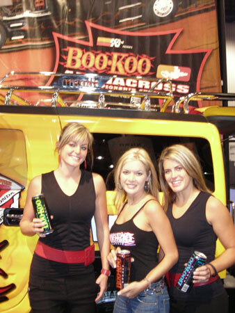 Three smiling women in black tops stand in front of a yellow vehicle and a Boo-Koo banner. Each holds a can of energy drink.