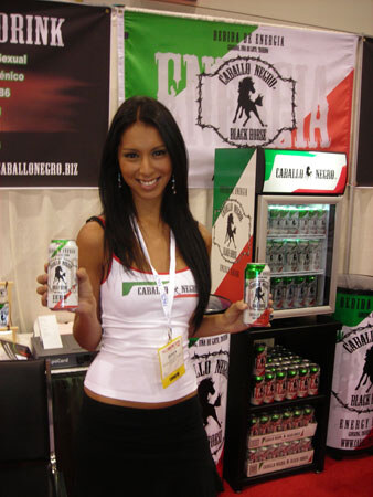 A smiling woman in a white top and badge holds two Caballo Negro energy drink cans beside a lit cooler and banners with a black horse logo.