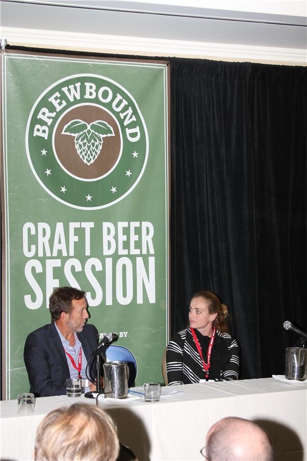 A man in a dark jacket and a woman in a striped top sit at a table in front of a green Brewbound Craft Beer Session banner, while audience members' heads are visible in the foreground.