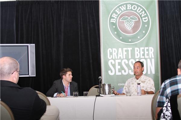 Two men sit at a draped table with a pitcher and microphone beside a green Brewbound Craft Beer Session banner, with audience members seated in the foreground.