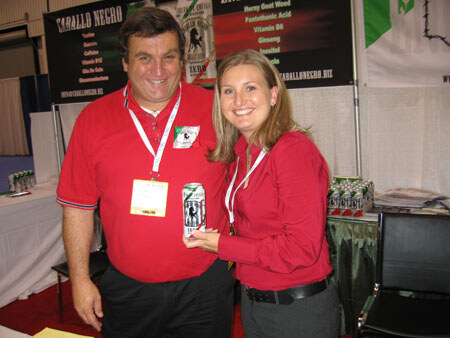 A man and a woman in red shirts and lanyards smile together at a booth with Caballo Negro banners behind them. The man holds a can of the drink.