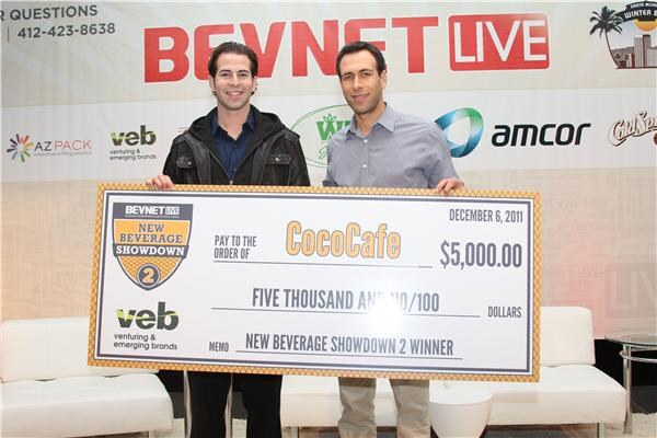 Two men hold a large check made out to CocoCafe for $5,000.00 marked New Beverage Showdown 2 Winner, in front of a BevNET LIVE sponsor backdrop.