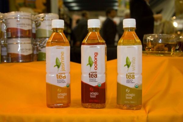 Three bottled organic teas from Adagio Teas, labeled jasmine, oolong and white, stand on an orange tablecloth. Jars of tea sit behind them and people are blurred in the background.
