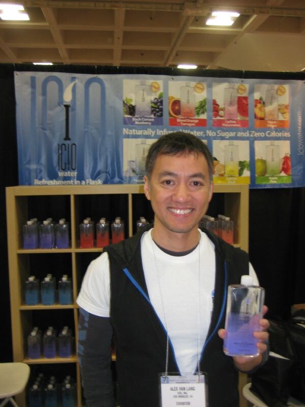 Alex Van Lang of ICIO, Inc. smiles and holds a flat bottle of purple-blue ICIO Water in front of shelves of bottles and an ICIO Water banner.
