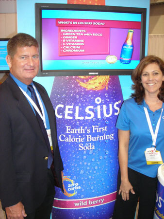 A man in a dark blazer and a woman in a blue polo, both wearing badges, stand beside a large Celsius wild berry bottle graphic beneath a Samsung screen listing ingredients.