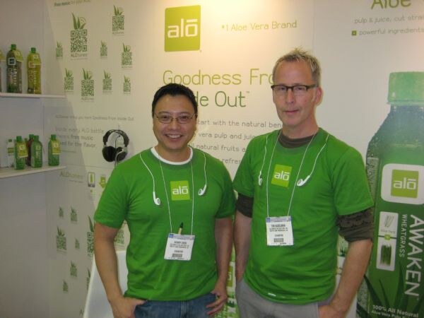 Two men in green alō T-shirts and earbuds around their necks stand in front of an alō display. Shelves of alō bottles and a large Awaken bottle graphic are behind them.