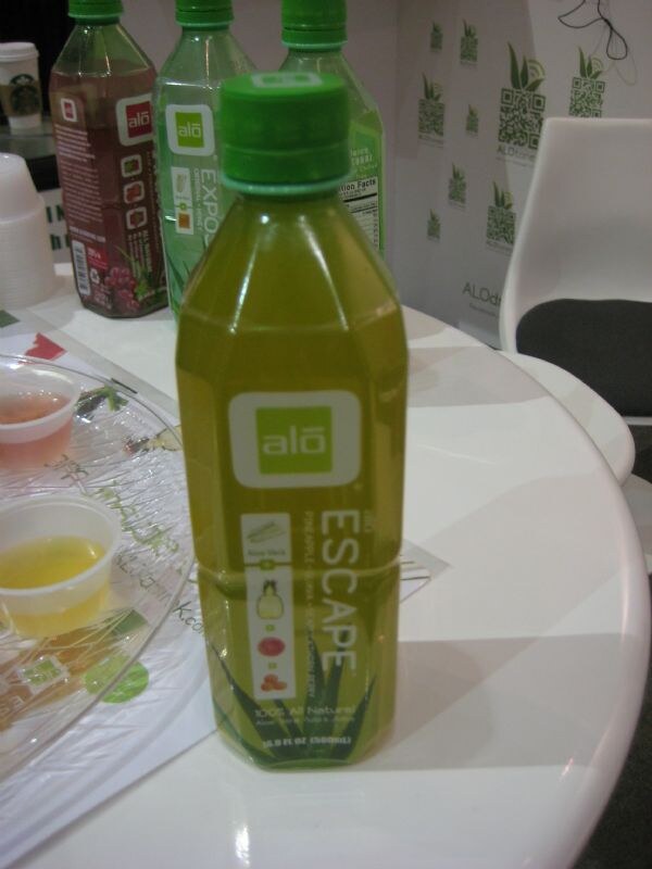 A green bottle of alō Escape aloe vera drink stands on a white table. More alō bottles, sample cups on a clear tray and a branded backdrop are behind it.