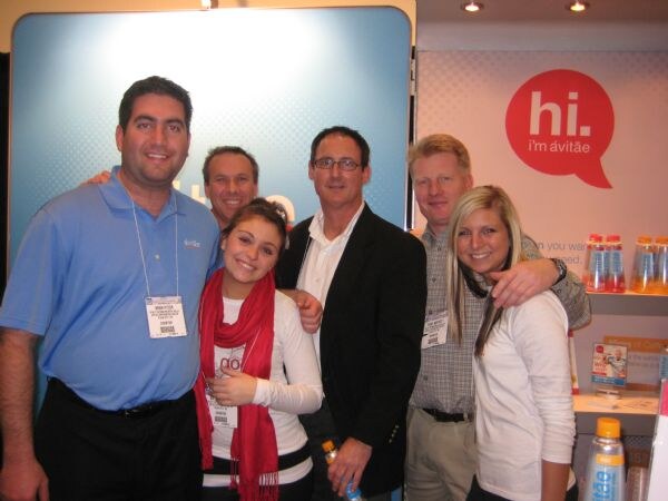 Five smiling people, three women and two men, pose close together in front of an ávitäe booth sign reading "hi. i'm ávitäe". Bottles of ávitäe water sit on a shelf and in the foreground.