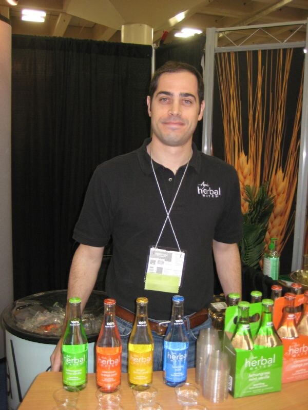 A man in a black Herbal Water polo and a badge stands behind a table of Ayala's Herbal Water bottles in green, orange, yellow and blue. Four-pack cartons and sample cups are beside them.