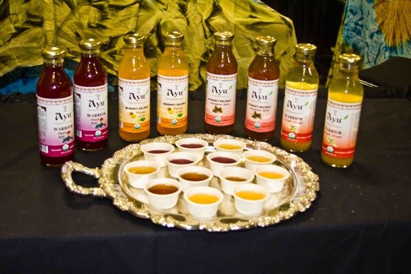 Eight bottles of Ayu herbal drinks, including Warrior and Woman's Balance varieties, stand behind a silver tray of small white sample cups on a black tablecloth.