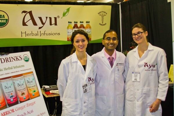 Two women and a man in white lab coats stand in front of an Ayu Herbal Infusions banner with Doctor's Drinks branding. A sign showing Ayu bottles is at the left.