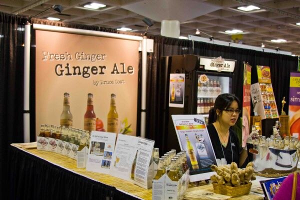 A woman with glasses sits behind the Bruce Cost Ginger Ale booth, which has a large Fresh Ginger Ale banner, a display cooler, bottle cartons, a basket of ginger and printed signs.