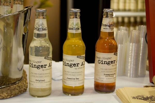 Three bottles of Bruce Cost Fresh Ginger Ale in Original, Jasmine Tea and Pomegranate flavors on a white table beside a metal ice bucket and a stack of clear plastic cups.