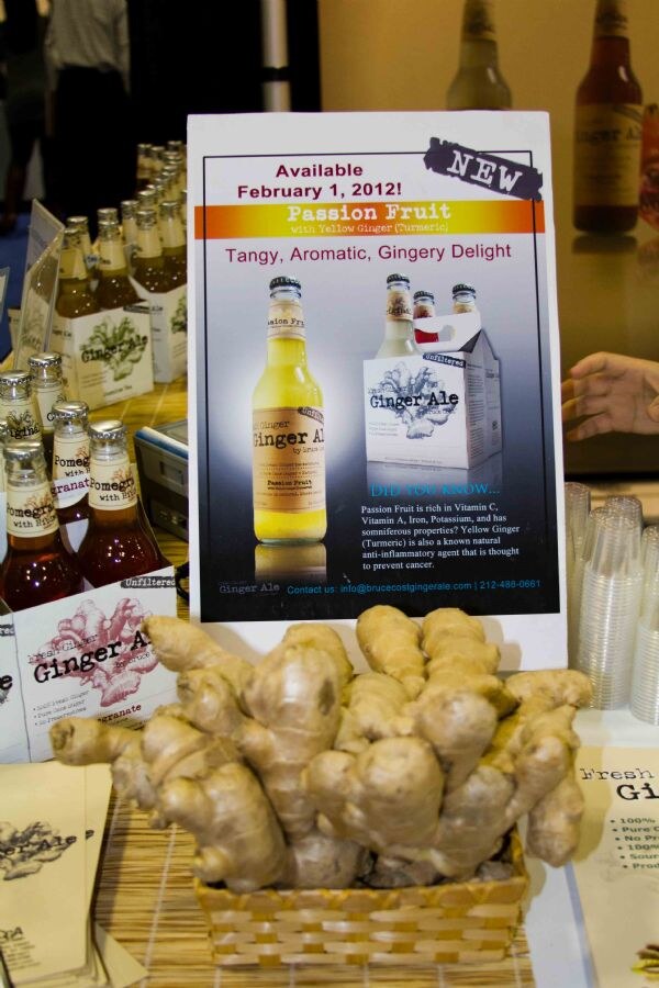 Booth sign for Bruce Cost Ginger Ale's new Passion Fruit flavor above a basket of fresh ginger root, with bottles, four-pack cartons and plastic cups around it.