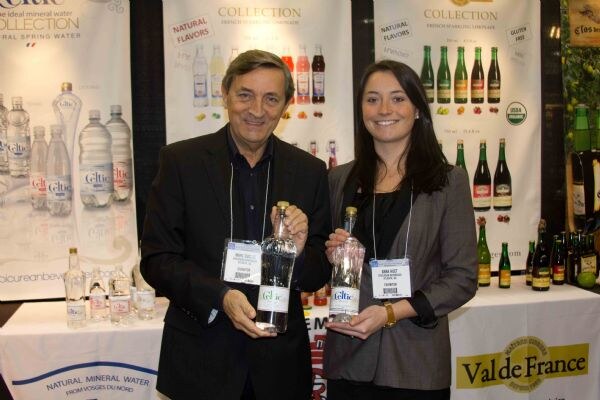 A man and a woman in dark jackets hold bottles of Celtic mineral water at a booth with Celtic water, Collection sparkling drinks and Val de France signs behind them.