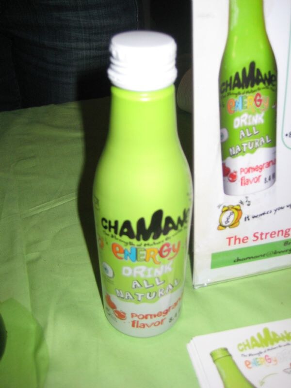 A green bottle of Chamane all natural energy drink, pomegranate flavor, stands on a green tablecloth next to a printed Chamane sign and a stack of flyers.