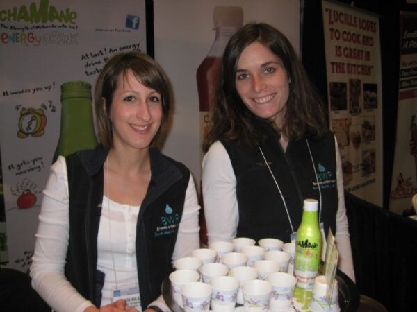 Two smiling women in dark vests stand behind a tray of small paper sample cups and a green Chamane energy drink bottle. Chamane banners are behind them.