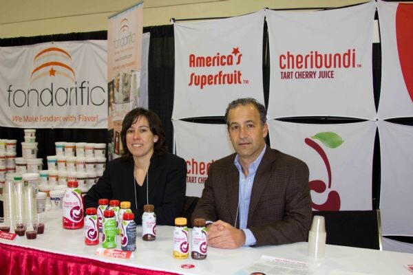 A woman in a black jacket and a man in a brown blazer sit at a table with Cheribundi tart cherry juice bottles and Fondarific fondant tubs, with Cheribundi and Fondarific banners behind them.