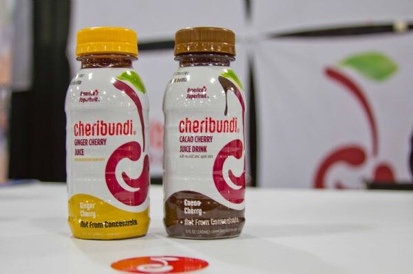 Two Cheribundi bottles on a white table: Ginger Cherry juice with a yellow cap and Cacao Cherry juice drink with a brown cap, in front of a cherry-logo banner.
