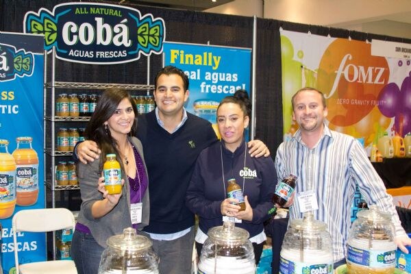 Four people smile and pose at the Cobá aguas frescas booth, two holding bottles, with Cobá banners, shelves of bottles and a FOMZ sign behind them.