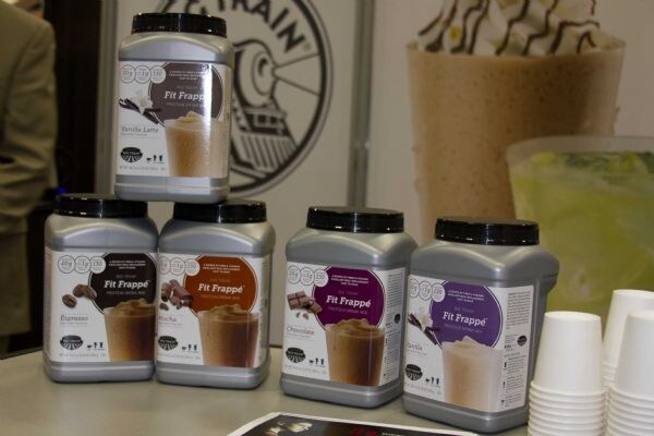 Four gray canisters of Fit Frappé mix stacked on a table: Vanilla Latte on top, Espresso, Mocha, Chocolate and Vanilla below. A blended frappé drink and stacked paper cups are behind them.