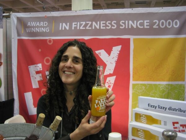 A smiling woman with long dark curly hair holds up a bottle of Fizzy Lizzy orange drink in front of a banner reading 'Award winning, in fizziness since 2000'. Boxes of bottles are at the right.