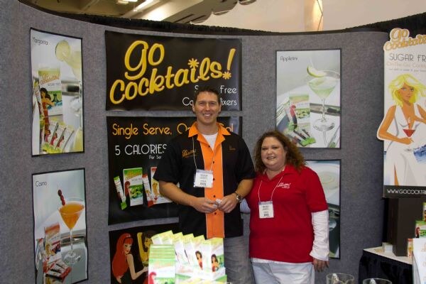 A man in a black and orange shirt and a woman in a red shirt, both wearing badges, stand in the Go Cocktails! booth. Cocktail photos and a 'Single Serve' sign hang on gray walls behind them.