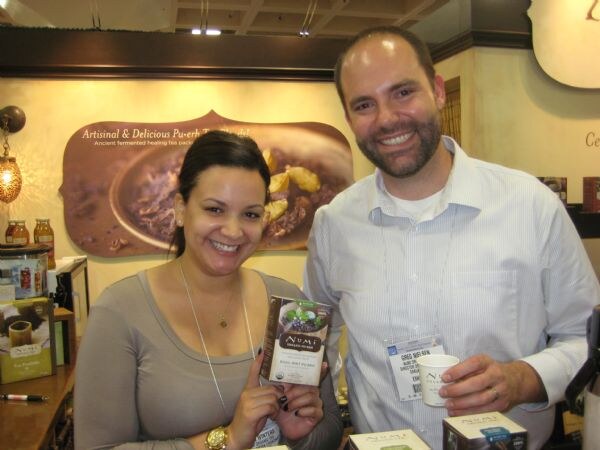 Greg Nelsen, at right, smiles and holds a paper cup beside a smiling woman holding a box of Numi tea, in front of a Numi display.