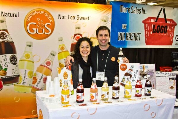 A smiling woman and man stand close together behind a table of GuS Grown-Up Soda bottles in several flavors. Large GuS banners with bottle graphics hang behind them.
