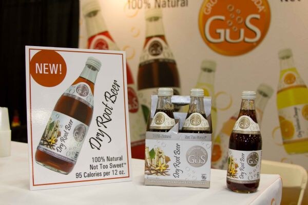 A sign reading 'New! Dry Root Beer' stands beside a four-pack and a single bottle of GuS Dry Root Beer on a table. A GuS banner with soda bottles is in the background.