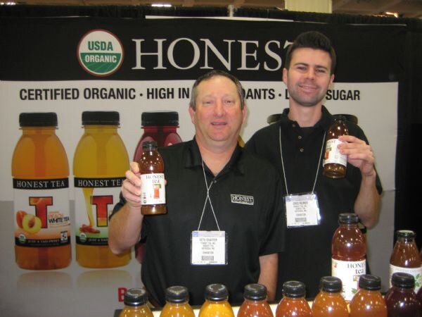 Two men in black shirts with exhibitor badges each hold a bottle of Honest Tea in front of a banner with organic tea bottles. More bottles line the table in the foreground.